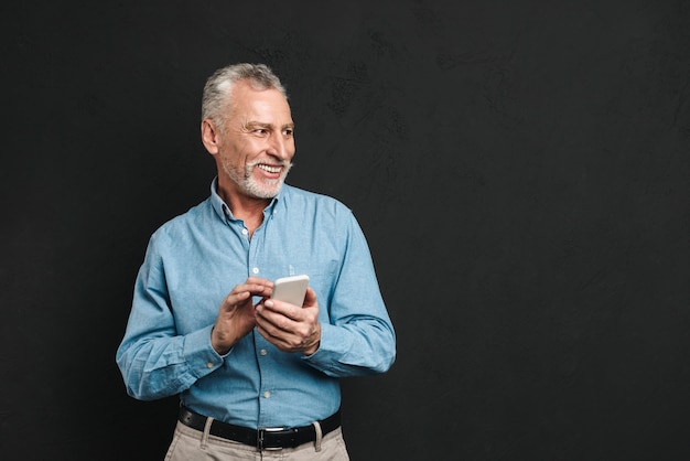 Immagine del pensionato maschio barbuto anni '60 con i capelli grigi in chat o navigazione internet sul cellulare, isolato su muro nero