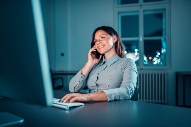 Immagine del primo piano della donna di affari sorridente che parla sul telefono mentre lavorando tardi alla notte.