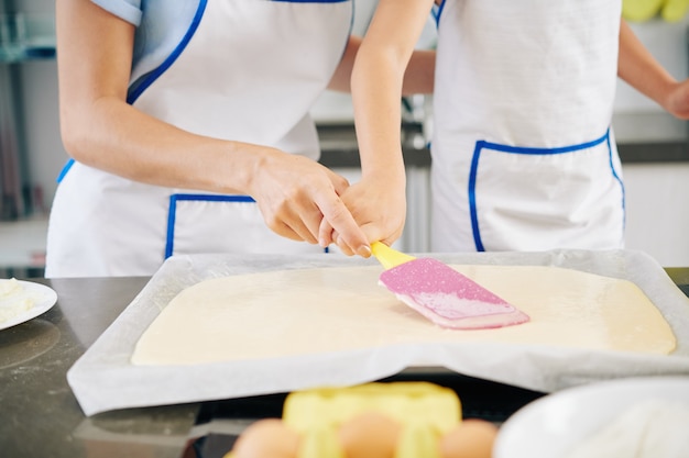 Immagine del primo piano della madre che aiuta la figlia a distribuire la pasta liquida nella teglia con una spatola di silicone