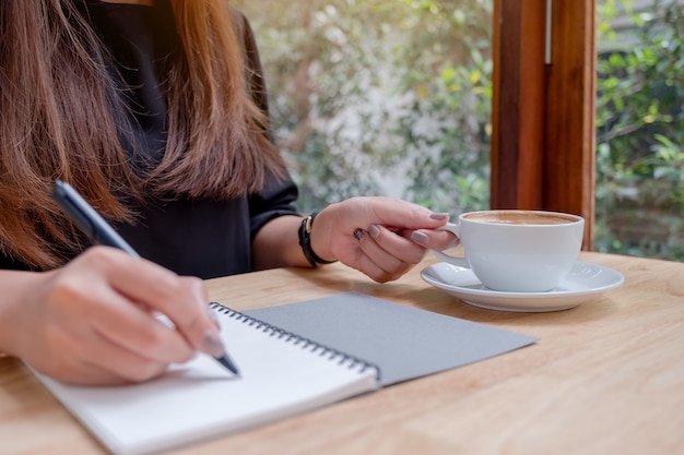 Immagine del primo piano della mano di una donna che annota su un taccuino in bianco bianco mentre bevendo caffè sulla tavola di legno