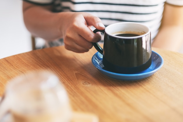 Immagine del primo piano della mano di una donna che tiene una tazza nera di caffè caldo sulla tavola di legno