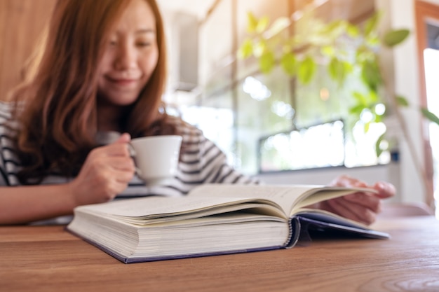Immagine del primo piano di bella donna asiatica che tiene e legge un libro mentre beve il caffè