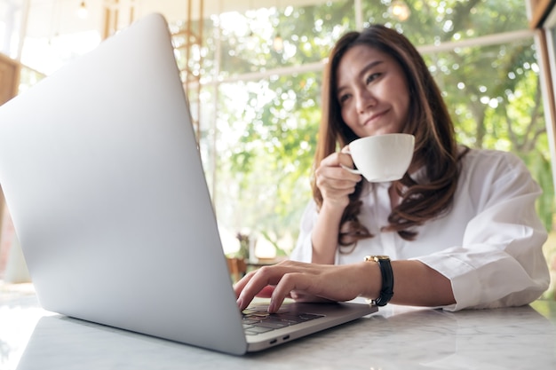 Immagine del primo piano di bella donna asiatica che utilizza computer portatile mentre bevendo caffè in caffè