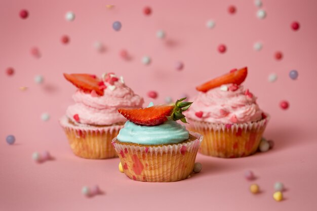 Immagine del primo piano di tre cupcakes colorati con fragole su superficie rosa con caramelle che cadono