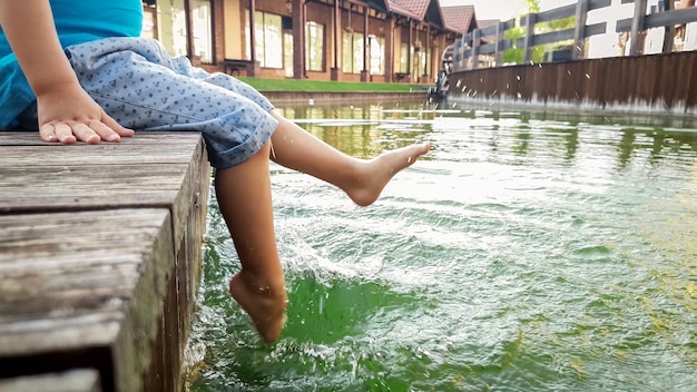 Immagine del primo piano di un bambino a piedi nudi seduto sul ponte di legno sul lago e che spruzza acqua con i piedi