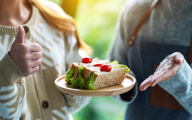 Immagine del primo piano di un cliente che fa il segno della mano del pollice mentre una cameriera che serve panino integrale nel piatto di legno