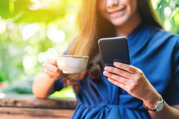 Immagine del primo piano di una bella donna asiatica che tiene e usa il telefono cellulare mentre beve caffè in giardino