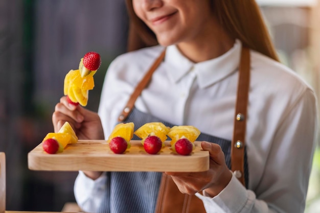 Immagine del primo piano di una bella donna chef che tiene frutta fresca mista su spiedini in un piatto di legno