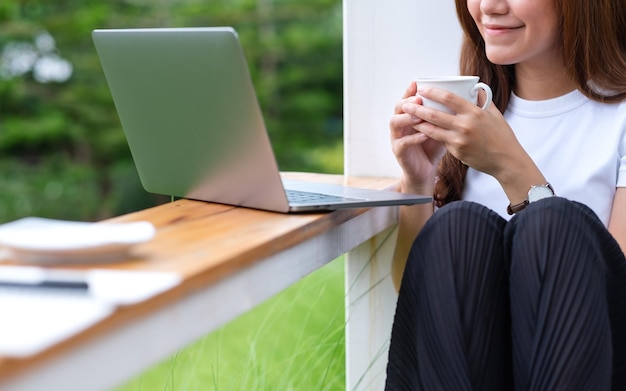 Immagine del primo piano di una bella giovane donna asiatica che beve caffè mentre si utilizza e si lavora al computer portatile in mezzo alla natura