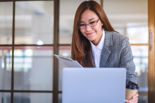 Immagine del primo piano di una bella giovane donna asiatica di affari che beve caffè e utilizza il computer portatile mentre lavora in ufficio