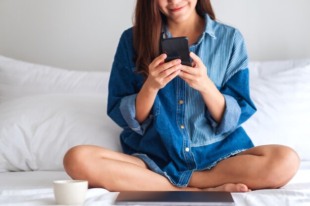 Immagine del primo piano di una bellissima giovane donna asiatica che usa il telefono cellulare e il laptop mentre è seduta su un letto bianco