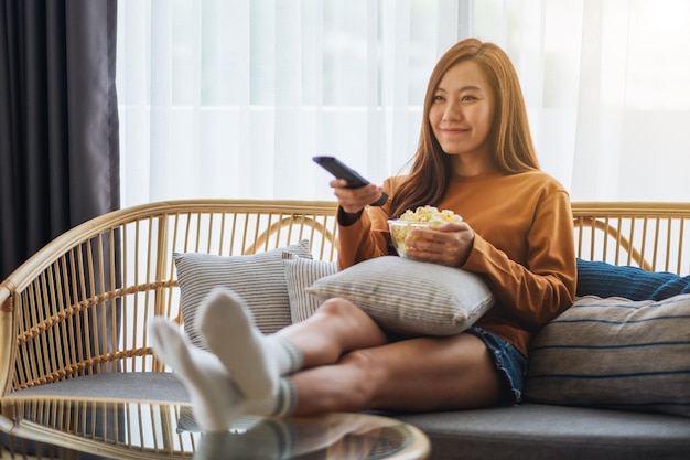 Immagine del primo piano di una bellissima giovane donna che mangia popcorn e cerca un canale con il telecomando per guardare la tv mentre è seduta sul divano di casa