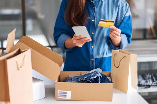 Immagine del primo piano di una bellissima giovane donna che utilizza il telefono cellulare e la carta di credito per lo shopping online con la borsa della spesa e la cassetta postale dei vestiti sul tavolo