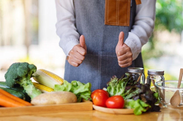 Immagine del primo piano di una chef donna che fa e mostra il pollice in alto segno della mano mentre prepara verdure miste fresche da cucinare in cucina