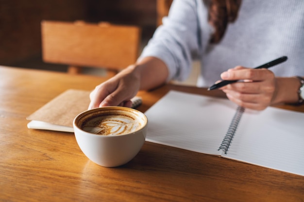 Immagine del primo piano di una donna che beve caffè e scrive su un taccuino in bianco sul tavolo