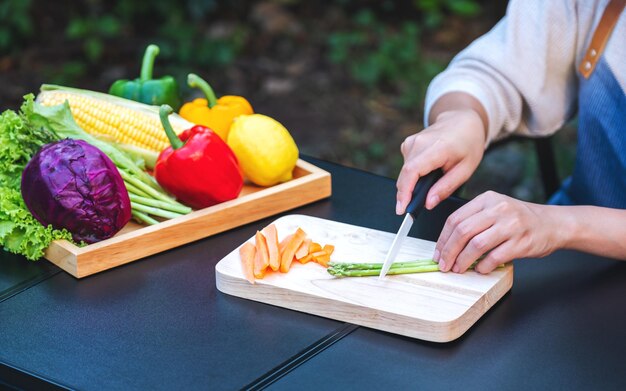 Immagine del primo piano di una donna che taglia e trita asparagi e carote con un coltello su una tavola di legno