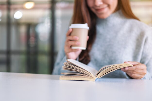 Immagine del primo piano di una donna che tiene e legge un libro mentre beve caffè su un tavolo di legno