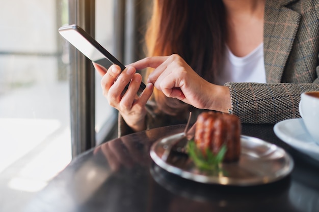 Immagine del primo piano di una donna che tiene e usa il telefono cellulare con una tazza di caffè e uno spuntino sul tavolo di un bar