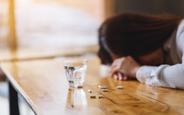 Immagine del primo piano di una donna malata che dorme sul tavolo con pillole bianche e un bicchiere d'acqua