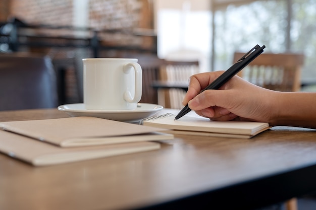 Immagine del primo piano di una mano che scrive sul taccuino in bianco con la tazza di caffè sul tavolo nella caffetteria