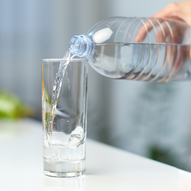 Immagine del primo piano di una mano femminile che tiene la bottiglia di acqua potabile e versando acqua nel bicchiere sul tavolo sullo sfondo della cucina.