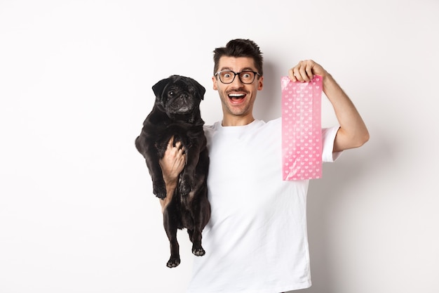 Immagine del proprietario di un animale domestico hipster, che tiene in mano un simpatico carlino nero e una borsa di cacca di cane, in piedi su sfondo bianco