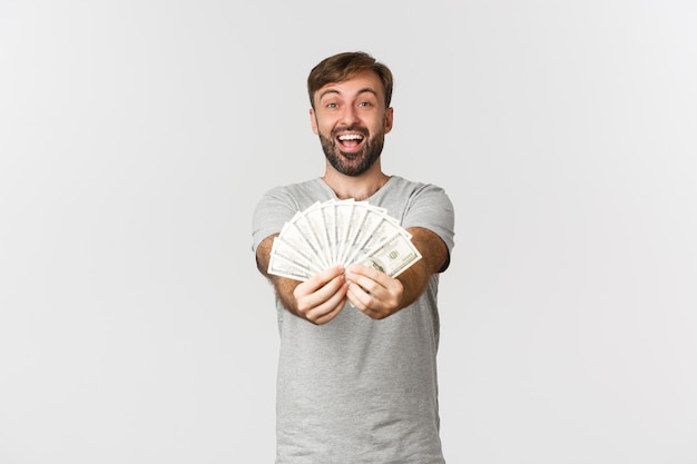 Immagine del ragazzo caucasico felice con la barba, dandoti soldi e sorridente, mostrando denaro, in piedi sopra