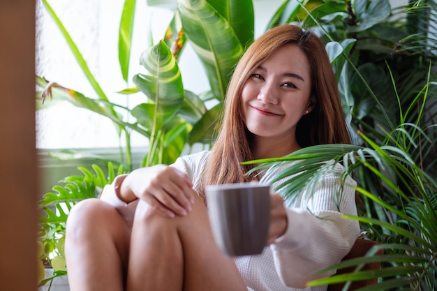 Immagine del ritratto di una bella donna asiatica che tiene e mostra una tazza di caffè mentre ci si rilassa sul balcone con il giardino delle piante d'appartamento a casa