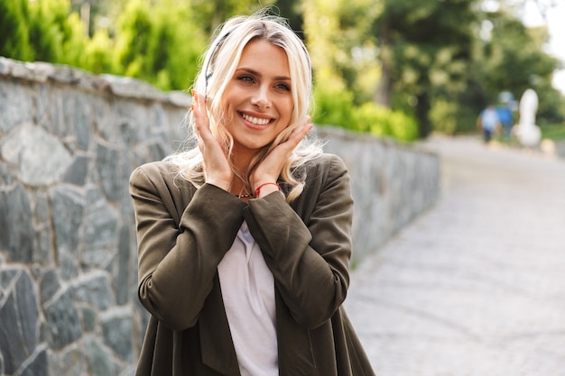 Immagine dell'affascinante donna 20s che indossa le cuffie sorridendo e ascoltando musica, mentre si cammina all'aperto