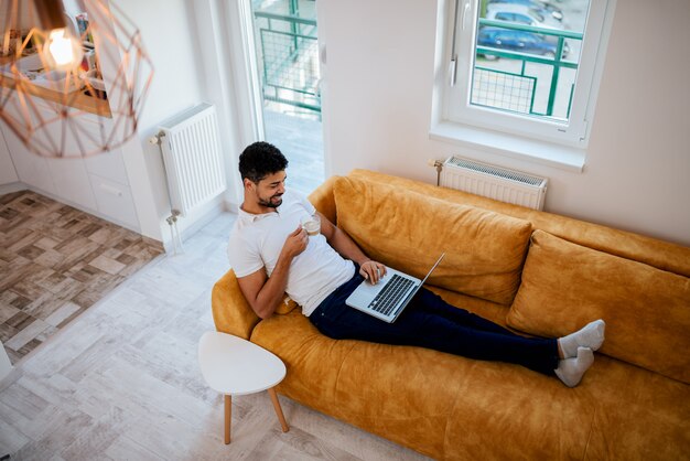 Immagine dell&#39;angolo alto dell&#39;uomo afroamericano che si rilassa sul sofà con il computer portatile e caffè.