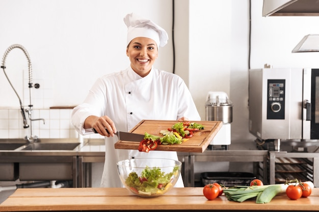 Immagine della bella donna capo che indossa l'uniforme bianca che produce insalata con verdure fresche, in cucina al ristorante