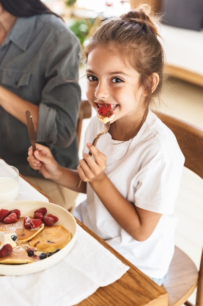 Immagine della bella madre di famiglia e della figlia piccola che si rallegrano e mangiano frittelle insieme mentre fanno colazione a casa al mattino