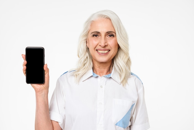 Immagine della donna adulta con capelli grigi che sorride e che fa pubblicità con la presentazione del telefono cellulare, isolata sopra la parete bianca