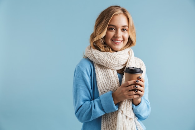 Immagine della donna bionda 20s avvolta in una sciarpa sorridente e tenendo il caffè da asporto