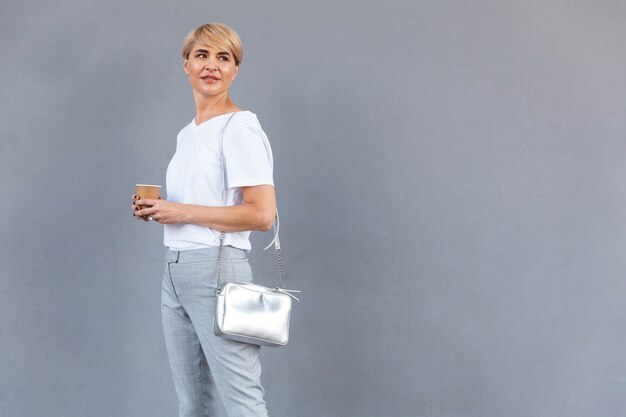 Immagine della donna bionda alla moda che indossa la maglietta bianca in piedi isolata sopra il muro grigio e guardando da parte copyspace con caffè da asporto