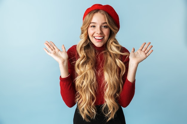 Immagine della donna bionda positiva 20s che indossa il berretto rosso che ride e che agita le mani mentre levandosi in piedi, isolata