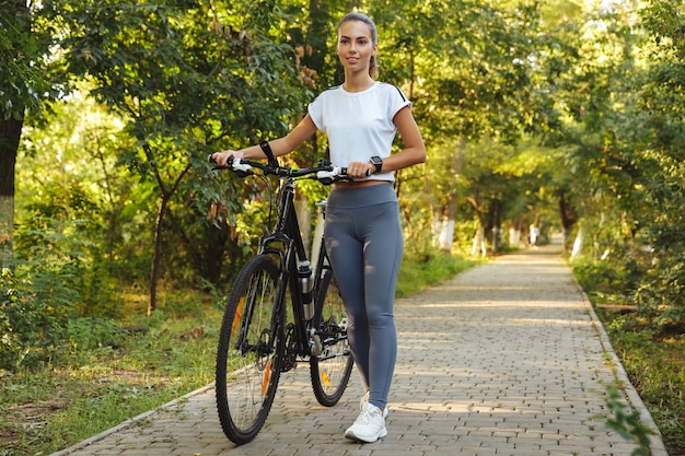 Immagine della donna felice 20s che cammina con la bicicletta attraverso il parco verde, durante la giornata di sole