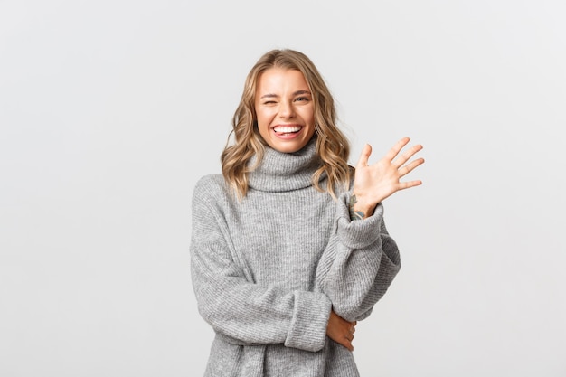 Immagine della donna felice amichevole in maglione grigio che dice ciao, sorridendo e agitando la mano per salutare la persona