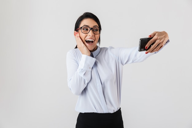 Immagine della donna graziosa dell'ufficio che indossa gli occhiali che prendono la foto del selfie sul telefono cellulare, isolato sopra il muro bianco