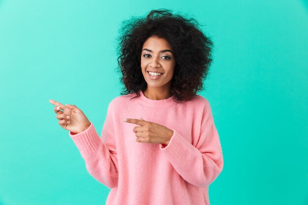 Immagine della donna riccia americana in camicia rosa e puntare il dito indice da parte su copyspace, isolato su muro blu