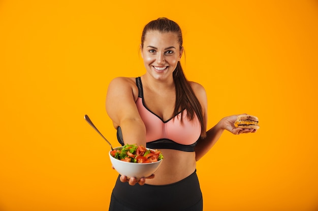 Immagine della donna sportiva in sovrappeso in tuta da ginnastica che mangia insalata verde dal piatto, isolata sopra priorità bassa gialla