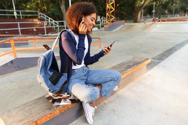 Immagine della giovane donna africana felice che si siede all'aperto nel parco utilizzando musica d'ascolto del telefono cellulare con gli auricolari.