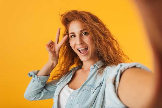 Immagine della giovane donna allegra sorridente e che mostra il segno di pace durante l'assunzione di foto selfie, isolato su sfondo giallo