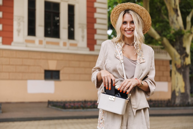 Immagine della giovane donna che indossa un abito e un cappello di paglia che mette il telefono cellulare nella sua borsa, mentre si cammina per la strada della città