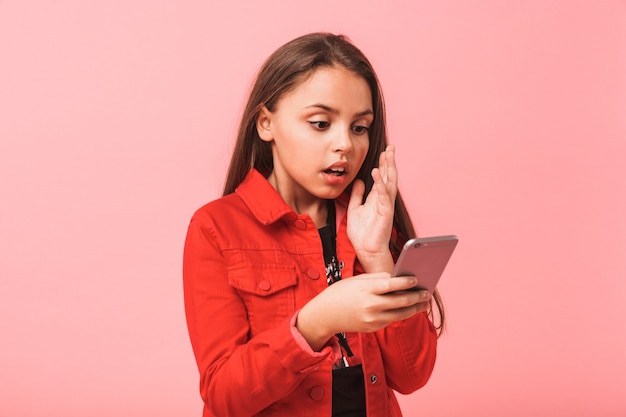 Immagine della ragazza adolescente sorpresa in casual utilizzando lo smartphone in piedi, isolata sopra la parete rossa