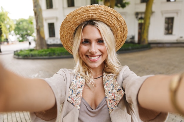 Immagine della signora caucasica che indossa tuta e cappello di paglia sorridente, tenendo selfie foto all'aperto nel centro cittadino