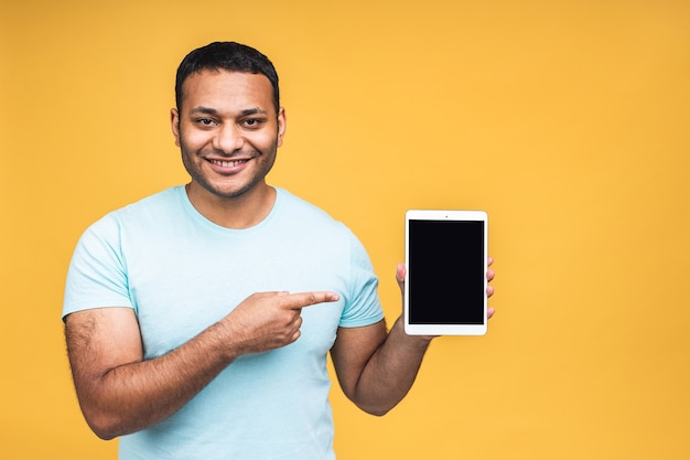 Immagine dello studio di un uomo nero indiano afroamericano positivo isolato su sfondo giallo che tiene tablet e lo mostra schermo vuoto con un sorriso felice come se consigliasse un prodotto o un servizio.