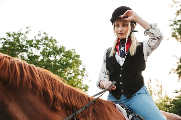 Immagine di adorabile giovane donna bionda che indossa un cappello a cavallo in cantiere in campagna