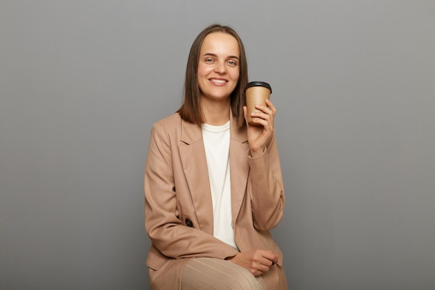 Immagine di attraente gioiosa donna allegra che indossa una giacca beige in posa isolata su sfondo grigio seduta con caffè da asporto nelle mani godendo di una bevanda calda