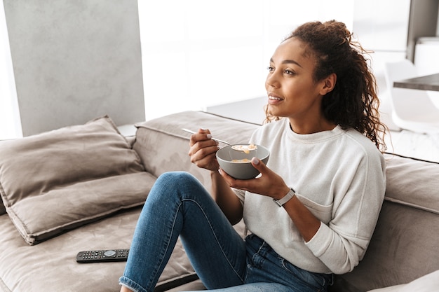 Immagine di bella donna afro-americana che mangia i fiocchi di mais con latte a colazione, mentre era seduto sul divano